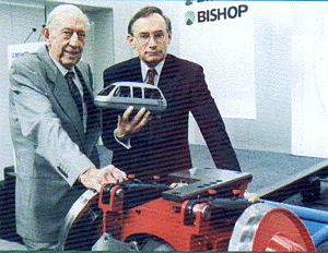 Photograph of two Bishop company figures holding a small Austrans vehicle model, with bogie hardware on a table in front of a BISHOP branded backdrop